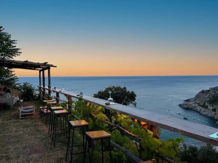 Un bar al aire libre con una larga barra de madera y taburetes en una terraza con vistas al mar al atardecer.