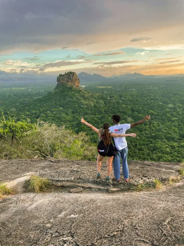 Two people from a WeRoad group trip embrace with open arms on a clifftop, overlooking a vast forest and a distant rock fortress.