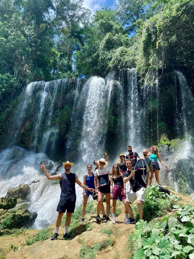 A WeRoad group trip posing for a photo on a rocky outcrop in front of a large, cascading waterfall in a lush forest.
