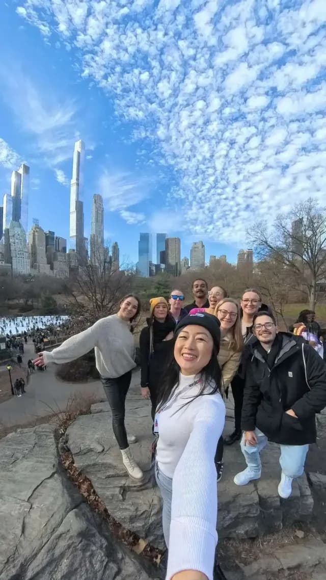 Una donna si scatta un selfie durante il suo viaggio di gruppo WeRoad, in posa su una grande roccia, con lo skyline di una città e una pista di pattinaggio sul ghiaccio sullo sfondo.