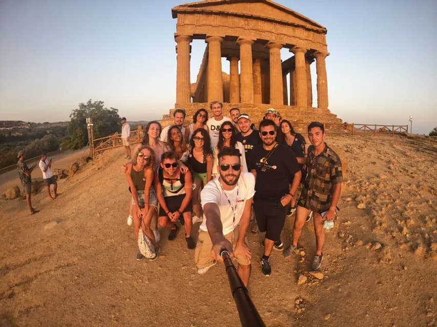 Un grupo de WeRoad se hace un selfi con un palo selfi frente a un antiguo templo griego en una colina polvorienta al atardecer.