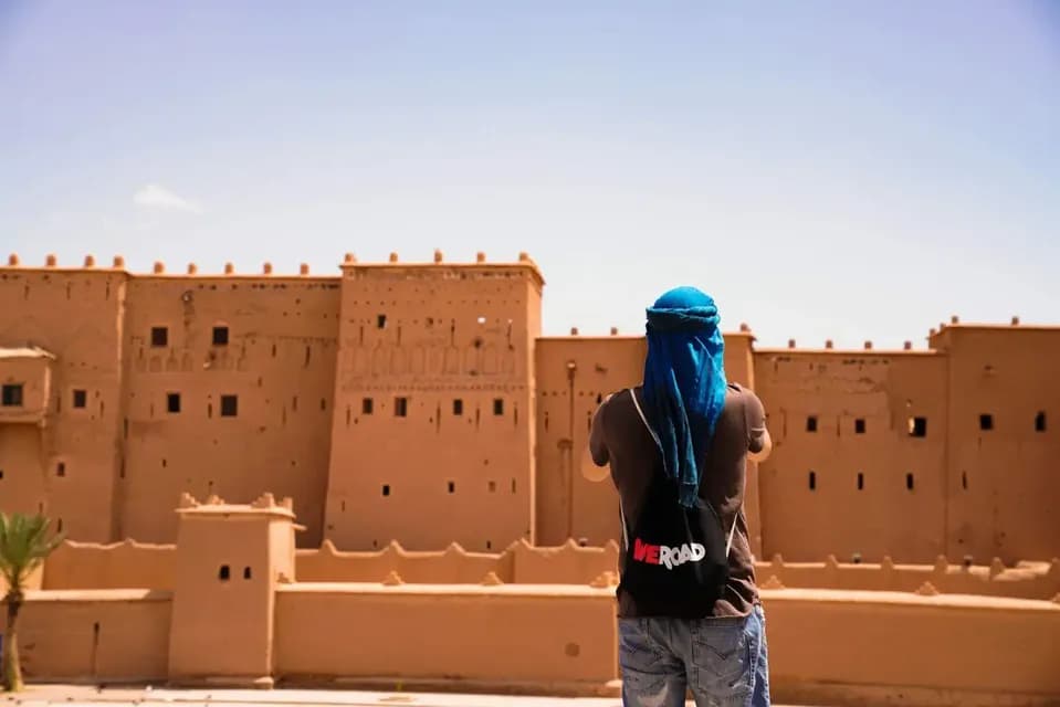 Una persona de espaldas, con un pañuelo azul y una mochila WeRoad, frente a una gran kasbah de color arena bajo un cielo despejado.