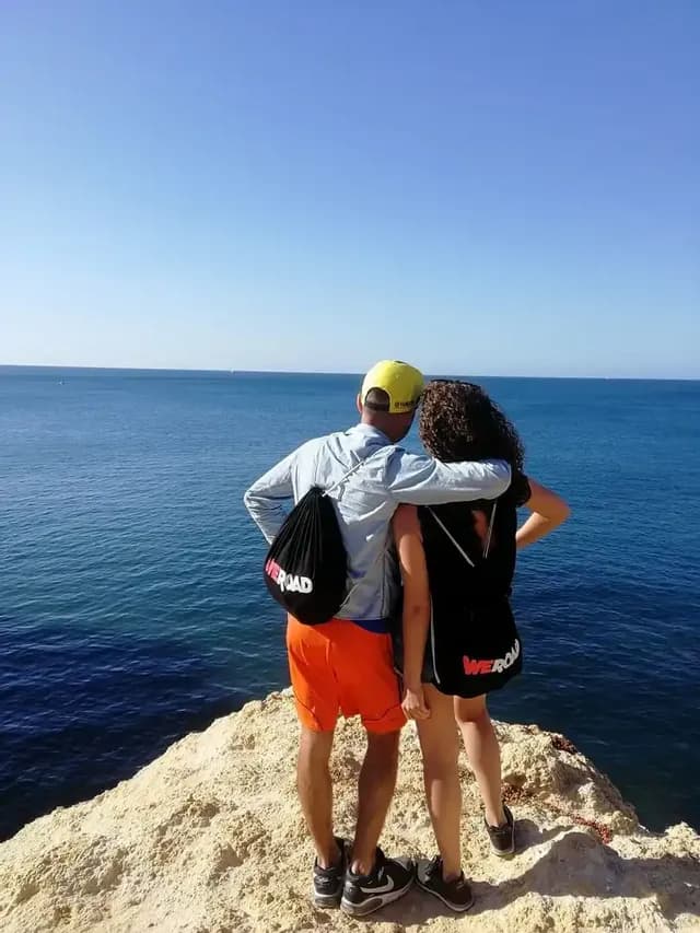 Un uomo di un viaggio di gruppo WeRoad con il braccio intorno a una donna, in piedi su una scogliera che si affaccia sull'oceano blu.