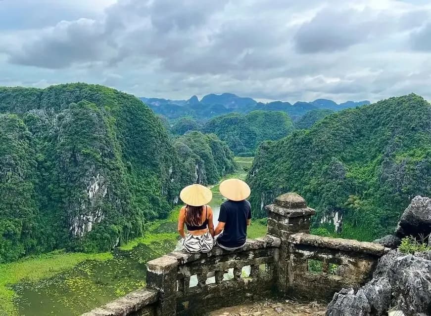 Un homme et une femme portant des chapeaux coniques sont assis sur un mur de pierre, contemplant une vallée fluviale verdoyante entourée de montagnes.