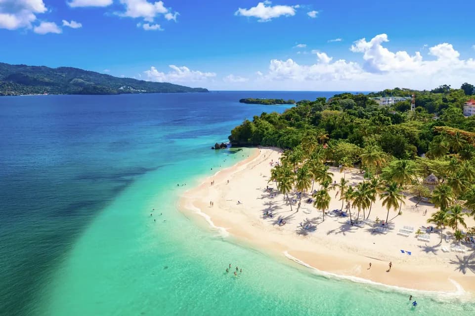 An aerial view of a tropical beach with people swimming in turquoise water and relaxing on white sand next to lush palm trees.