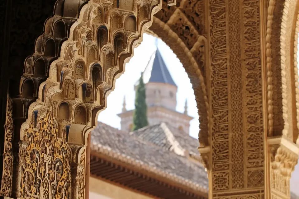 Un primer plano de un arco morisco ornamentado con intrincadas tallas, enmarcando una vista de una torre distante con techo de tejas.
