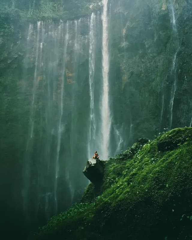 Una persona siede su una grande roccia su un pendio coperto di muschio, con una cascata alta e nebbiosa sullo sfondo.