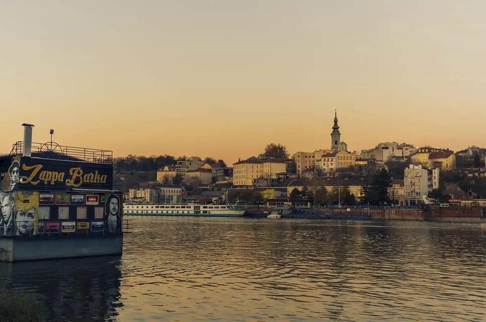 Una casa flotante llamada Zappa Barka flota en un río con el horizonte de una ciudad, incluyendo un campanario de iglesia, visible al fondo al atardecer.