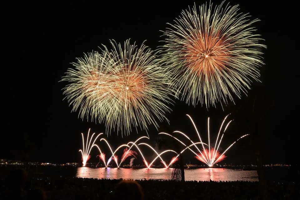 Fuochi d'artificio dorati e rossi esplodono sull'acqua di notte mentre una folla osserva dalla riva.