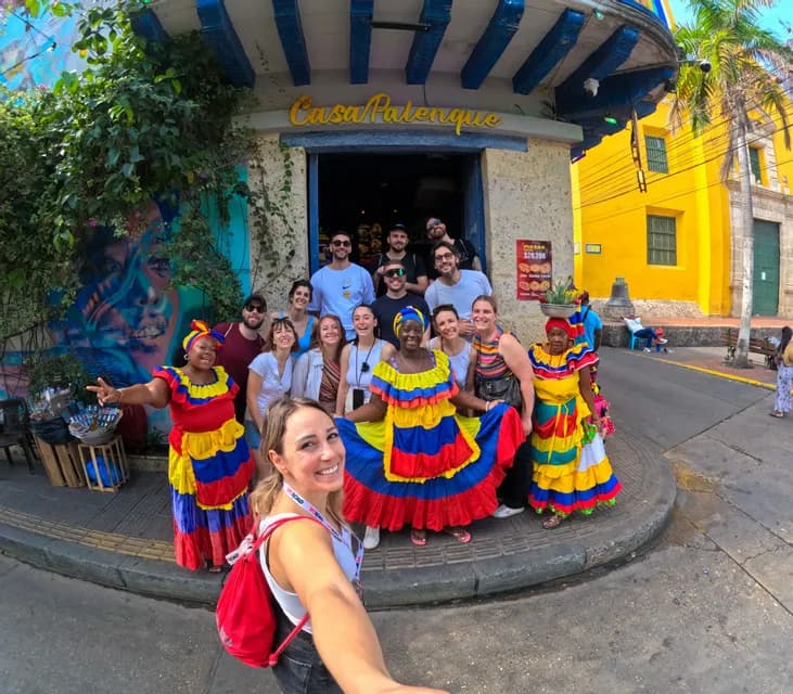 A WeRoad group trip poses for a selfie with two women in traditional colorful dresses on a city street.