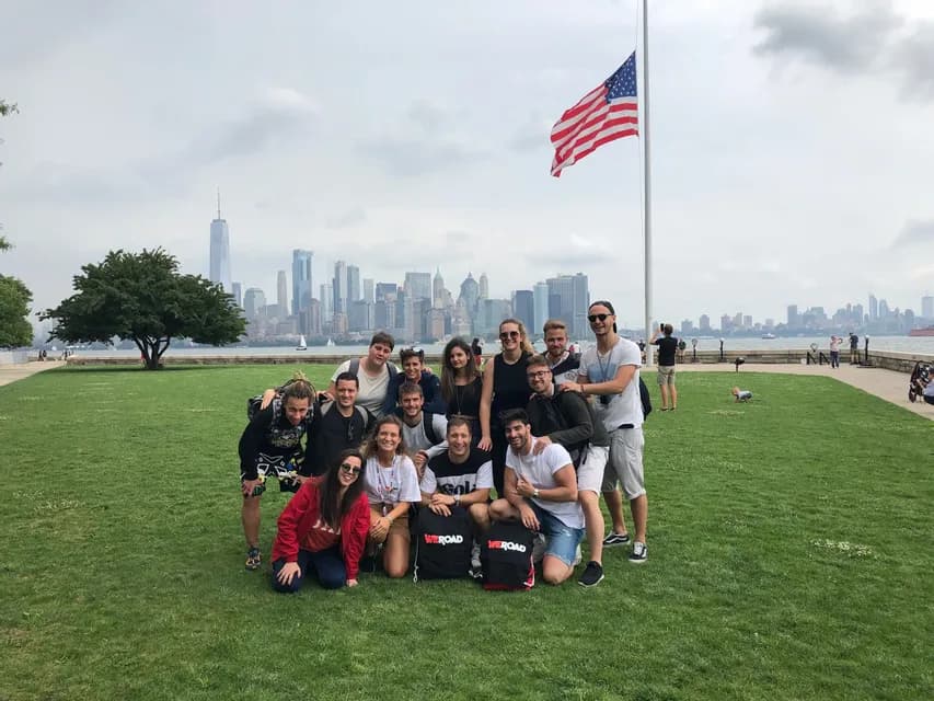 Un groupe WeRoad prend la pose pour une photo sur une pelouse verte, avec un drapeau américain et la skyline d'une ville en arrière-plan.