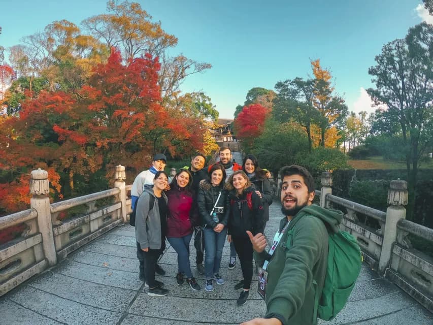 Eine WeRoad Gruppe macht lächelnd ein Selfie auf einer Steinbrücke, umgeben von bunten Herbstbäumen.