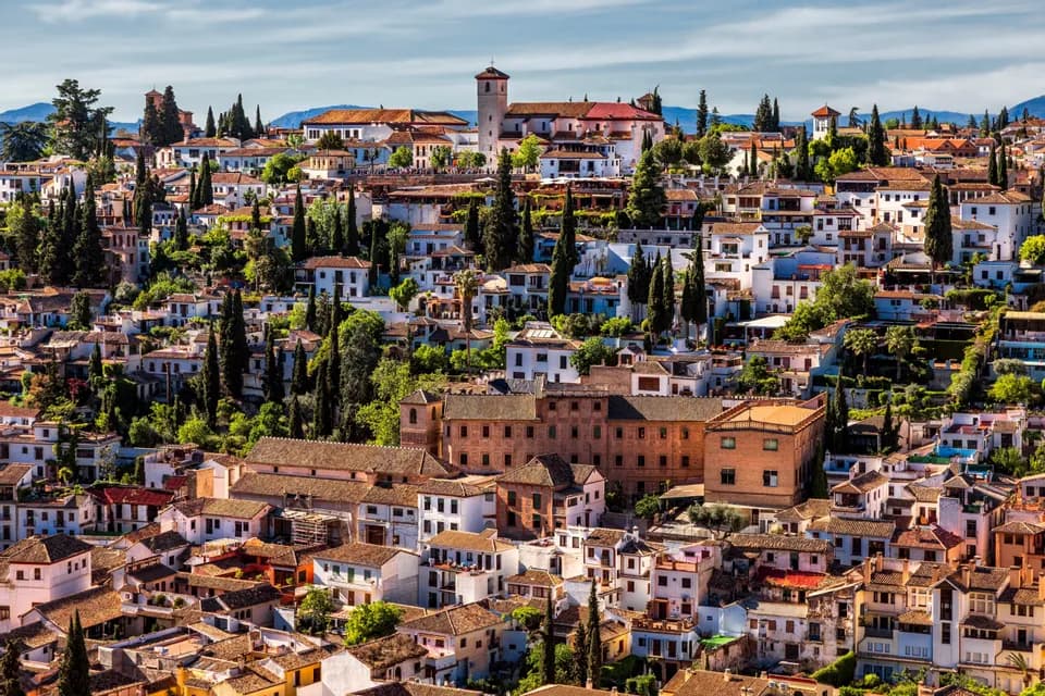 Una fitta città collinare con edifici bianchi, tetti in tegole di terracotta e alti alberi verdi sotto un cielo parzialmente nuvoloso.