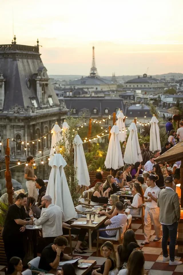 Persone socializzano ai tavoli di un affollato bar panoramico al tramonto, con la Torre Eiffel visibile nello skyline della città alle loro spalle.