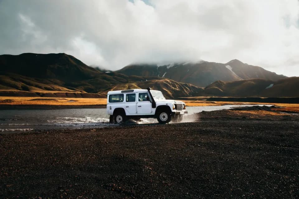 Un véhicule tout-terrain blanc traverse une rivière peu profonde dans un paysage montagneux avec des sommets enneigés sous un ciel nuageux.