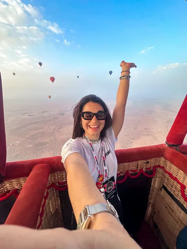 A woman on a WeRoad group trip smiles for a selfie in a hot air balloon basket, flying above a desert with other balloons.