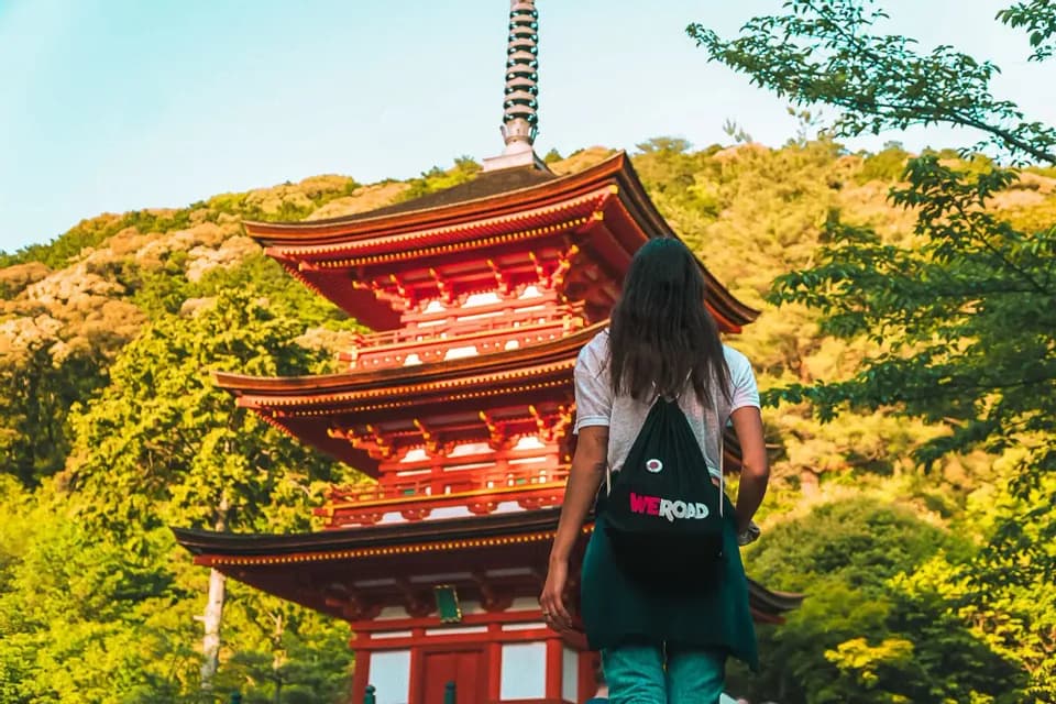 A woman from behind, wearing a WeRoad backpack, looks at a traditional multi-tiered red pagoda surrounded by green trees.