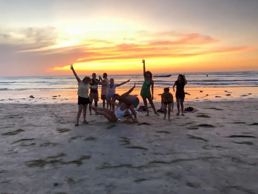 A WeRoad group trip of people posing playfully on a sandy beach with the ocean behind them during a colorful sunset.