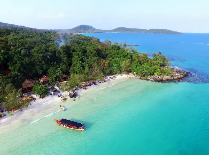 Vista aerea di una barca a coda lunga ancorata in acque cristalline turchesi, vicino a una spiaggia di sabbia bianca fiancheggiata da una fitta foresta.