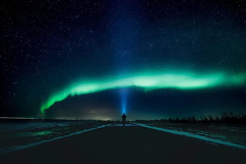 A person stands on a road at night, shining a flashlight up at the green aurora borealis in a starry sky.