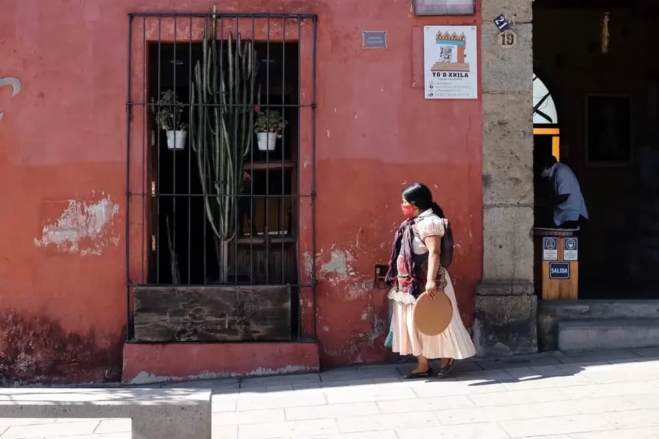 Una donna in abito tradizionale e una mascherina cammina lungo un marciapiede accanto a un edificio rosso-arancione con una finestra inferriata.