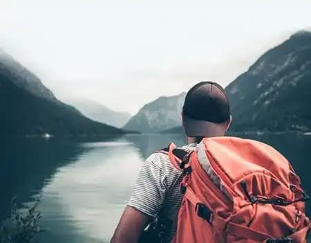 Una persona con uno zaino arancione e un cappellino osserva un lago calmo circondato da montagne nebbiose.