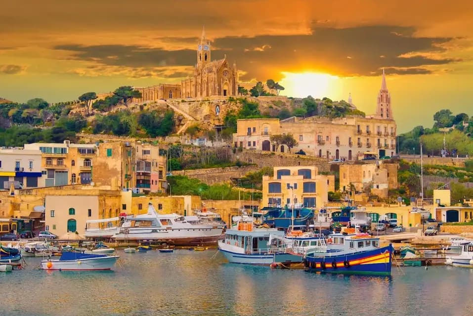 A coastal town with a church on a hill overlooks a harbor full of boats and yachts at sunset.