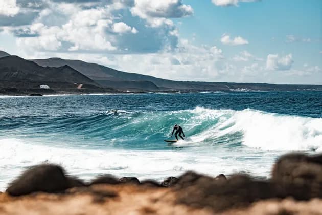 Un surfista in muta nera cavalca un'onda turchese, con una costa rocciosa e montuosa sullo sfondo.