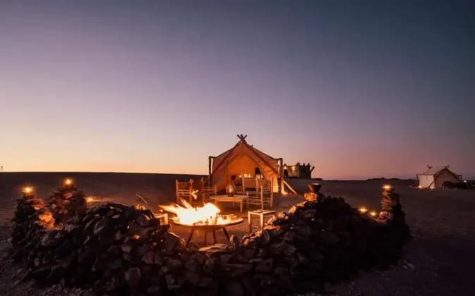 A campfire burns in a rock-lined pit in front of a large, illuminated tent in a desert campsite at dusk.