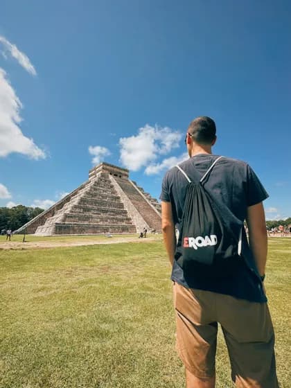 Ein Mann von hinten gesehen mit einem WeRoad-Rucksack, der eine große steinerne Stufenpyramide auf einem grasbewachsenen Feld unter blauem Himmel betrachtet.
