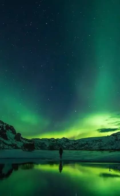Una persona de pie en una orilla nevada, observando la aurora boreal verde reflejarse en el agua bajo un cielo estrellado.