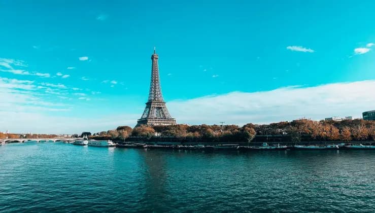 La Torre Eiffel se alza frente a un ancho río bordeado de árboles otoñales y barcos amarrados, bajo un cielo azul brillante.