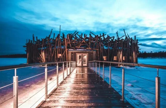 Una passerella in legno illuminata conduce a un edificio moderno con un'unica facciata a doghe, immerso in un paesaggio innevato al tramonto.