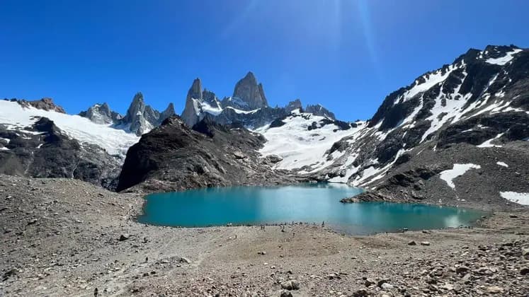 Un lago alpino turchese circondato da una riva rocciosa e montagne frastagliate e innevate sotto un cielo limpido e luminoso.