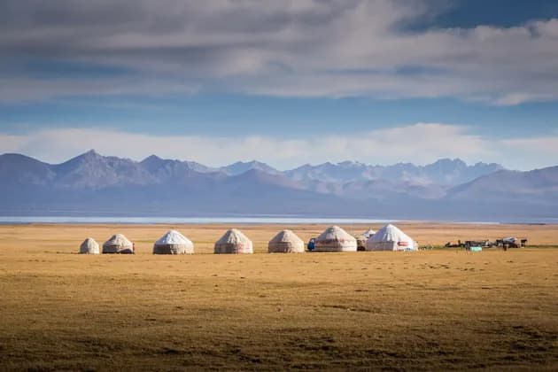 Une rangée de yourtes dans une plaine herbeuse, face à un lac et une chaîne de montagnes lointaine.