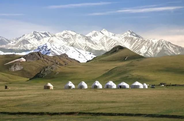 A row of yurts sits on a wide green plain, with rolling hills and a vast, snow-capped mountain range behind them.