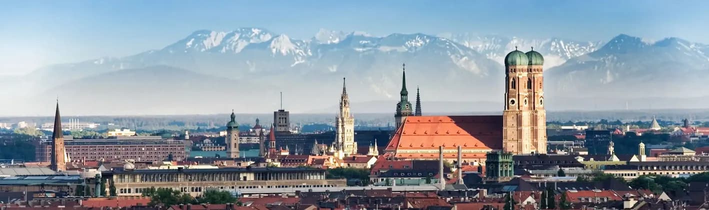 Un paesaggio urbano panoramico con tetti rossi e campanili, e una grande catena montuosa innevata sullo sfondo.
