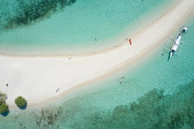 Una vista aerea di una lingua di sabbia bianca in acqua turchese, dove persone nuotano vicino a un kayak e una barca a bilanciere ormeggiata.