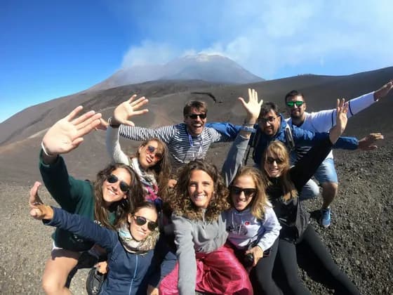 Un grupo de WeRoad se toma un selfi en una ladera rocosa, con un volcán humeante al fondo bajo un cielo azul claro.