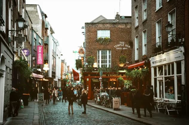 Una folla di persone su una strada di ciottoli di fronte a The Temple Bar, un pub storico in mattoni decorato con luci.