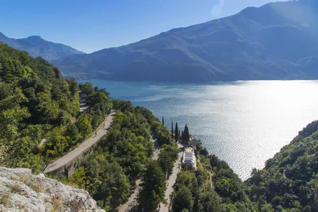 Una strada tortuosa su un verde versante di montagna domina un grande lago soleggiato e lontane catene montuose.