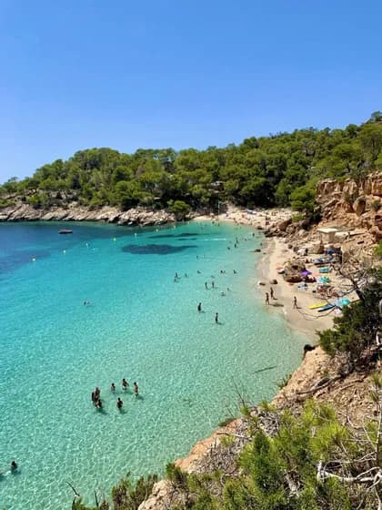 Una vista dall'alto di una cala con una spiaggia sabbiosa, dove le persone nuotano nelle acque limpide e turchesi sotto un cielo azzurro brillante.