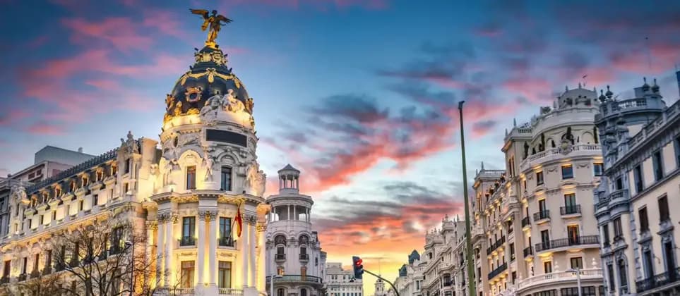 Edifici cittadini decorati, uno con tetto a cupola e statua dorata, sono illuminati contro un cielo vibrante al tramonto.