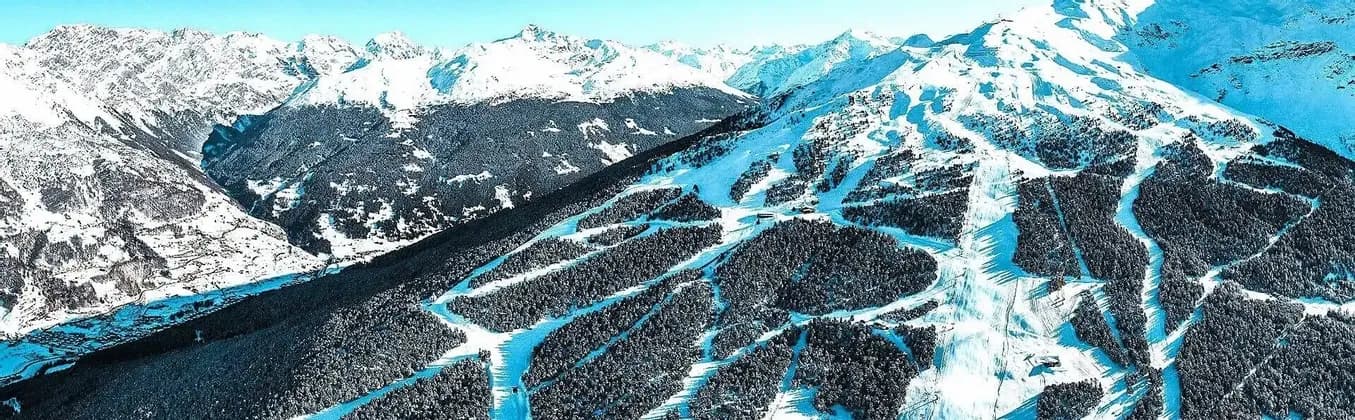 Veduta aerea di una catena montuosa innevata con piste da sci scavate tra fitte foreste sotto un cielo sereno.