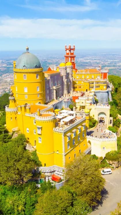 Una vista aerea di un grande castello colorato con torri gialle e rosse, situato su una collina verde e alberata sotto un cielo azzurro e limpido.