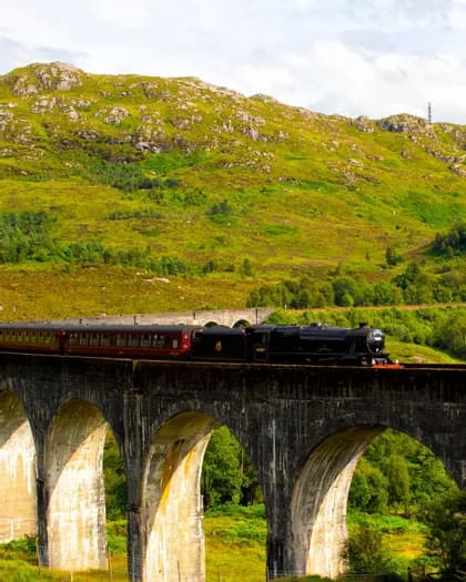 Un treno a vapore nero con carrozze rosse attraversa un viadotto in pietra, con una rigogliosa montagna verde sullo sfondo.