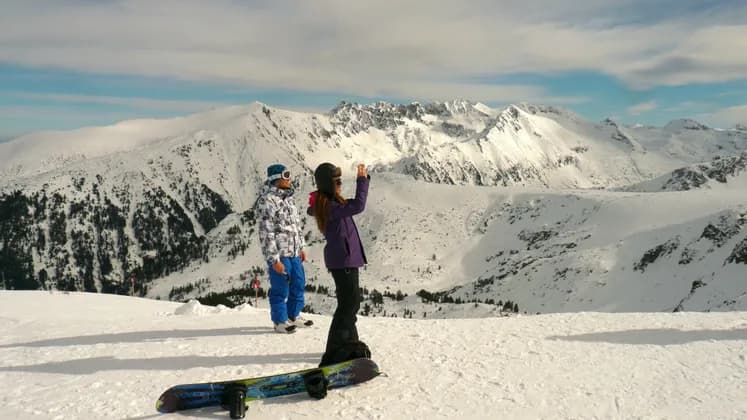 Zwei Snowboarder stehen auf einem verschneiten Berggipfel, einer fotografiert die weite, schneebedeckte Bergkette.