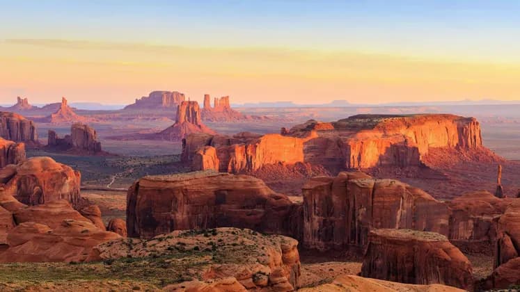 Eine weite Wüstenlandschaft mit großen roten Felsformationen und Tafelbergen, beleuchtet vom warmen Licht einer aufgehenden oder untergehenden Sonne.