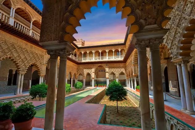 Un cortile in stile moresco riccamente decorato con una piscina centrale e giardini, visto attraverso un grande arco a conchiglia contro un cielo al tramonto.