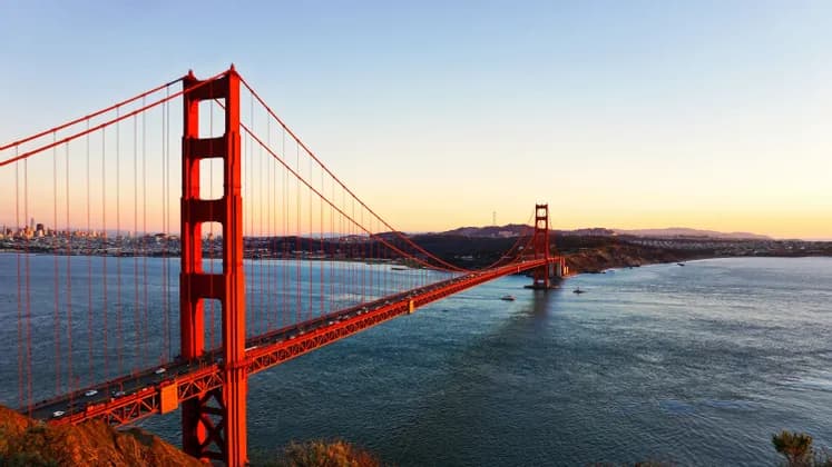 A large red suspension bridge spanning a bay at sunset, with cars crossing it and a city skyline in the background.